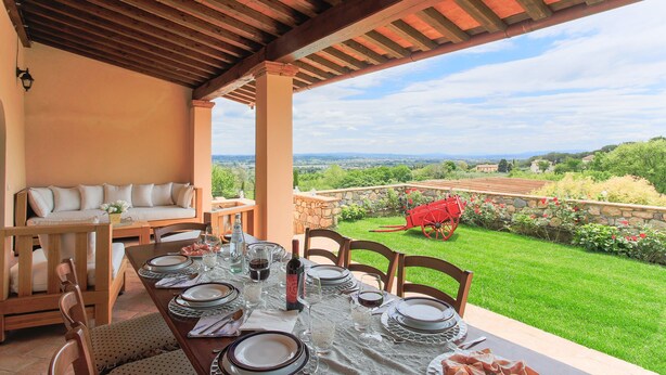 An outdoor dining area with a table set for four, a sofa, and a view of a lush garden and distant landscape.