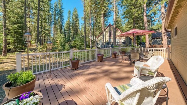 A wooden deck with outdoor furniture, surrounded by tall pine trees, and a building with a red roof in the background. - Sequoia Pines - 411 Sequoia Circle (Plumas Eureka)