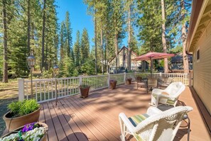 A wooden deck with outdoor furniture, surrounded by tall pine trees, and a building with a red roof in the background. - Sequoia Pines - 411 Sequoia Circle (Plumas Eureka)