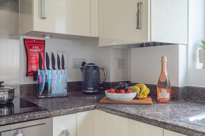 Et køkken med en bordplade i granit, kaffemaskine, en flaske Prosecco, en skål med frugt og et sæt knive.