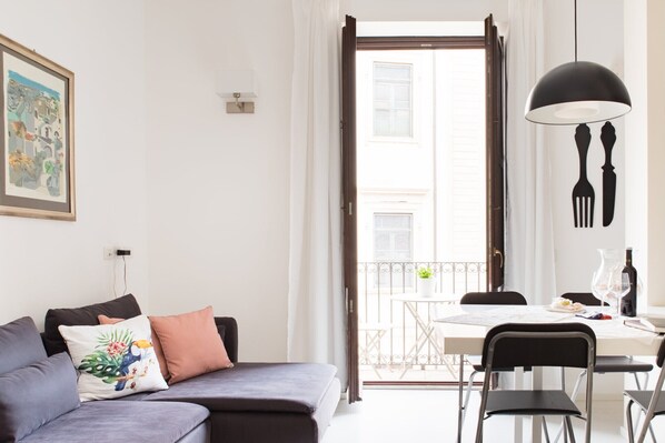 Un salon moderne comprenant un canapé, une table à manger avec des chaises, un balcon offrant une vue sur les immeubles et un luminaire suspendu.