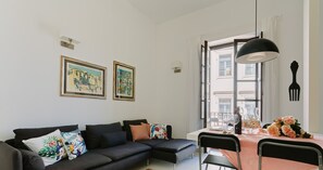 Un salon moderne avec un canapé d’angle noir, une table à manger recouverte d’une nappe couleur pêche et une grande fenêtre donnant sur des immeubles.