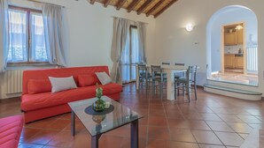 A spacious living area with a red sofa, a glass coffee table, and a dining table with chairs. There is a kitchen visible through an archway.
