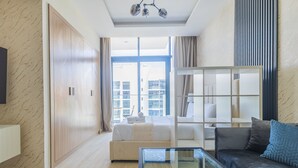 A modern hotel room with a large bed, a glass coffee table, a sofa, and a view of the city through large windows. - Nasma Luxury Stays - Riviera 10 (Dubai)