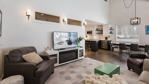 A modern living room with a large flat-screen TV, a sofa, a coffee table, and a kitchen area in the background. - The Honeycomb | AC, Pool, Hot Tub, Golf & Ski (Durango)