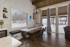 Ein moderner Essbereich mit einem langen Holztisch, vier Stühlen, einer Pendelleuchte und einem großen Fenster mit Jalousien.