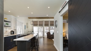 A modern kitchen with a central island, dark cabinetry, and a dining area with a large window. - The Honeycomb | AC, Pool, Hot Tub, Golf & Ski (Durango)