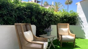 Twee rieten stoelen met witte kussens, een klein tafeltje en een bloempot op een terras met groen kunstgras en een witte muur.