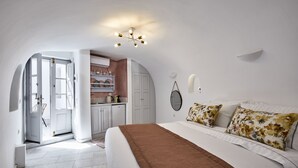 A bedroom with a large bed, a brown throw blanket, and floral pillows. There is a kitchen area visible through an open door. - El Verano Hideaway Villa (Santorini)