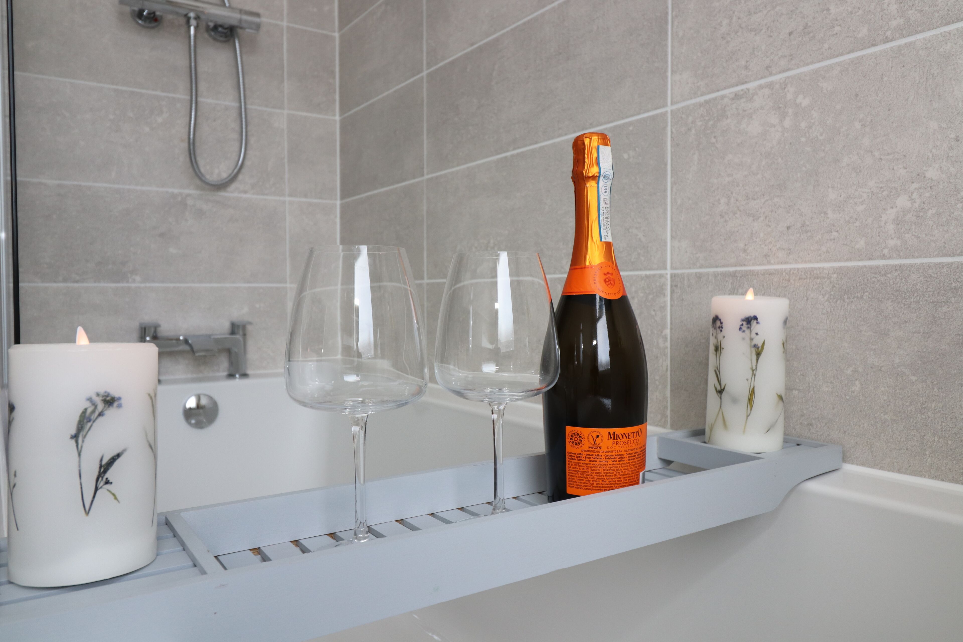 A bathroom with a bathtub, a shower, and a shelf with a bottle of wine, glasses, and candles.