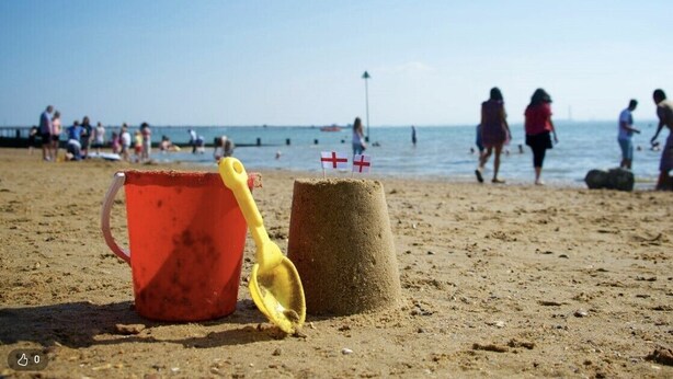 Una platja sorrenca amb un cub vermell i una pala groga, un castell de sorra amb dues banderes, i persones al fons.