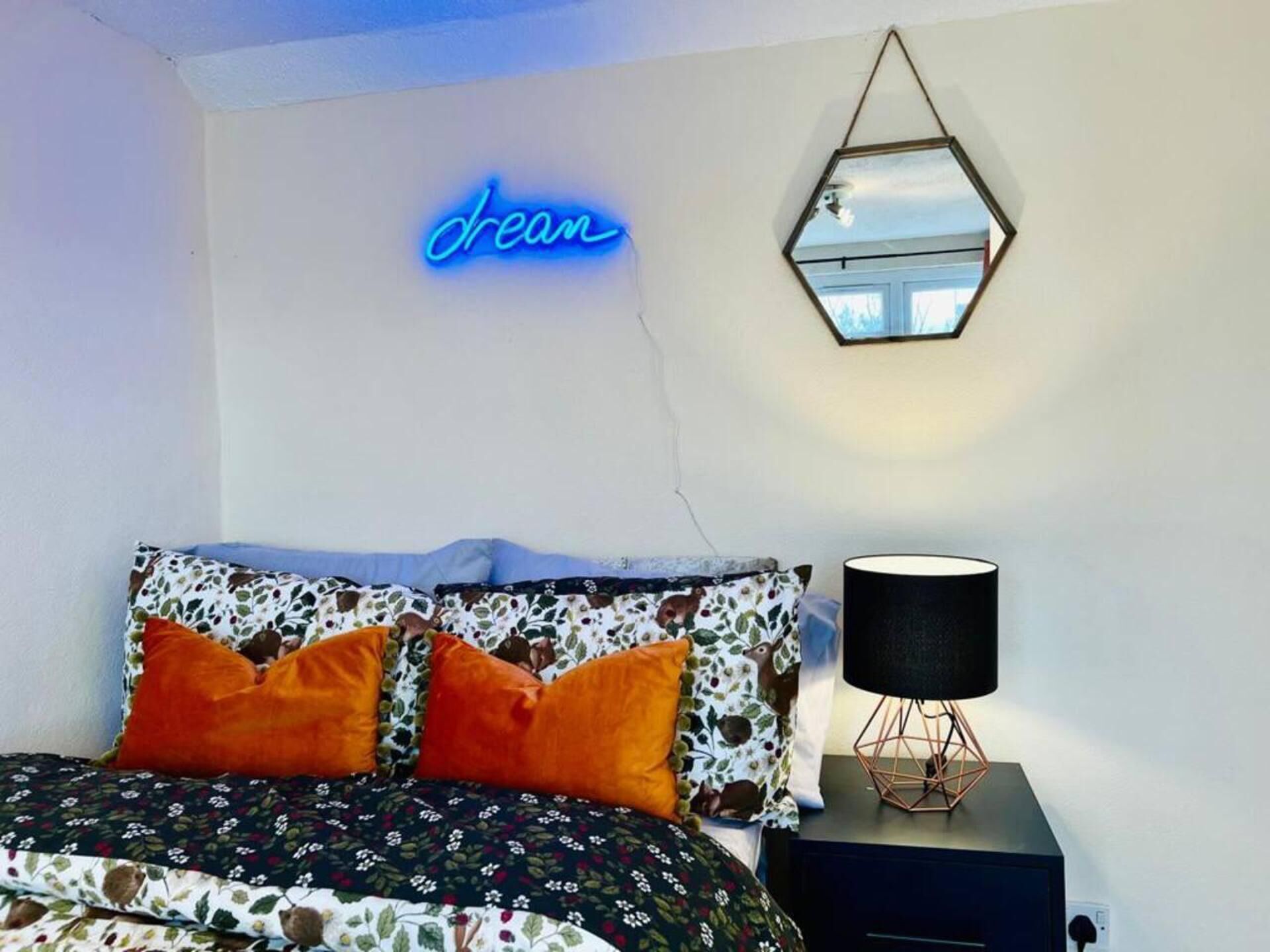 A bedroom with a bed, a neon 'dream' sign, a geometric mirror, and a lamp on a bedside table.