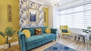 A living room with a teal sofa, yellow and red cushions, a yellow armchair, a chandelier, and a large window with blinds.