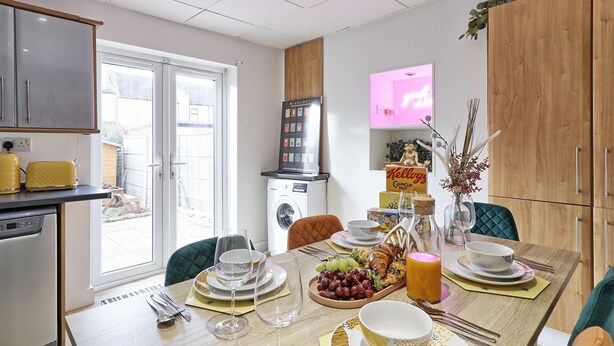 A dining area with a table set for two, a washing machine, and a television in the background.