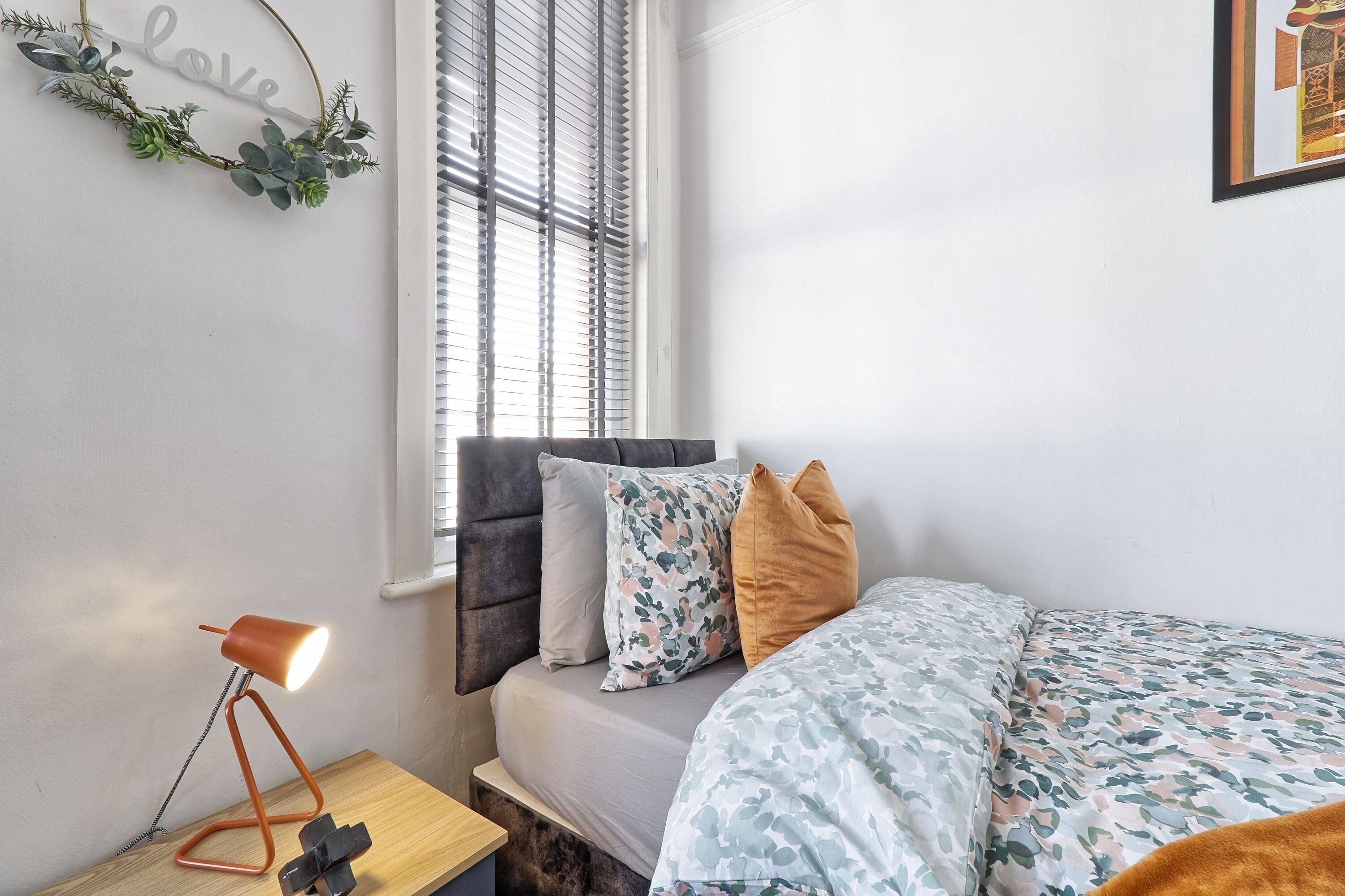 Ein Schlafzimmer mit Bett, Schreibtisch mit Lampe, Fenster mit Jalousien und Wanddekoration.