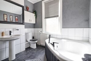 A modern bathroom with a white sink, toilet, and bathtub. There is a window with blinds, a mirror, and a plant on the wall.