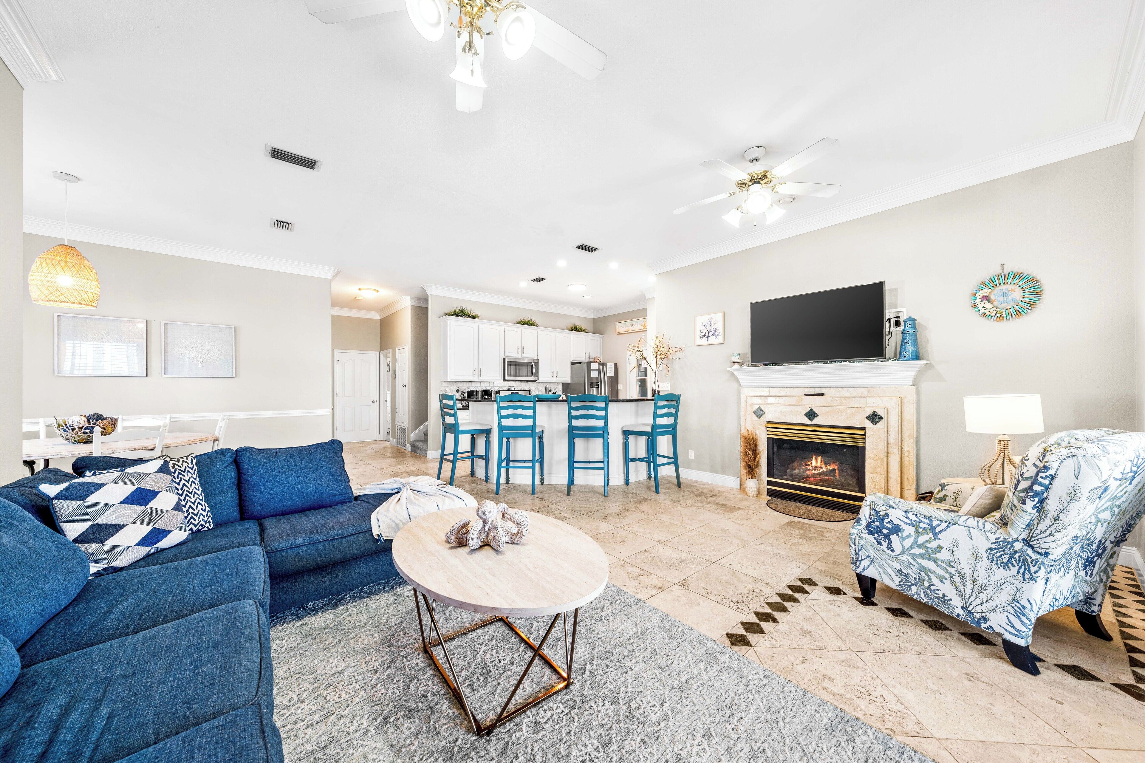 A modern living room with a sectional sofa, a round coffee table, and a fireplace. There is a kitchen area with blue chairs and a dining table.