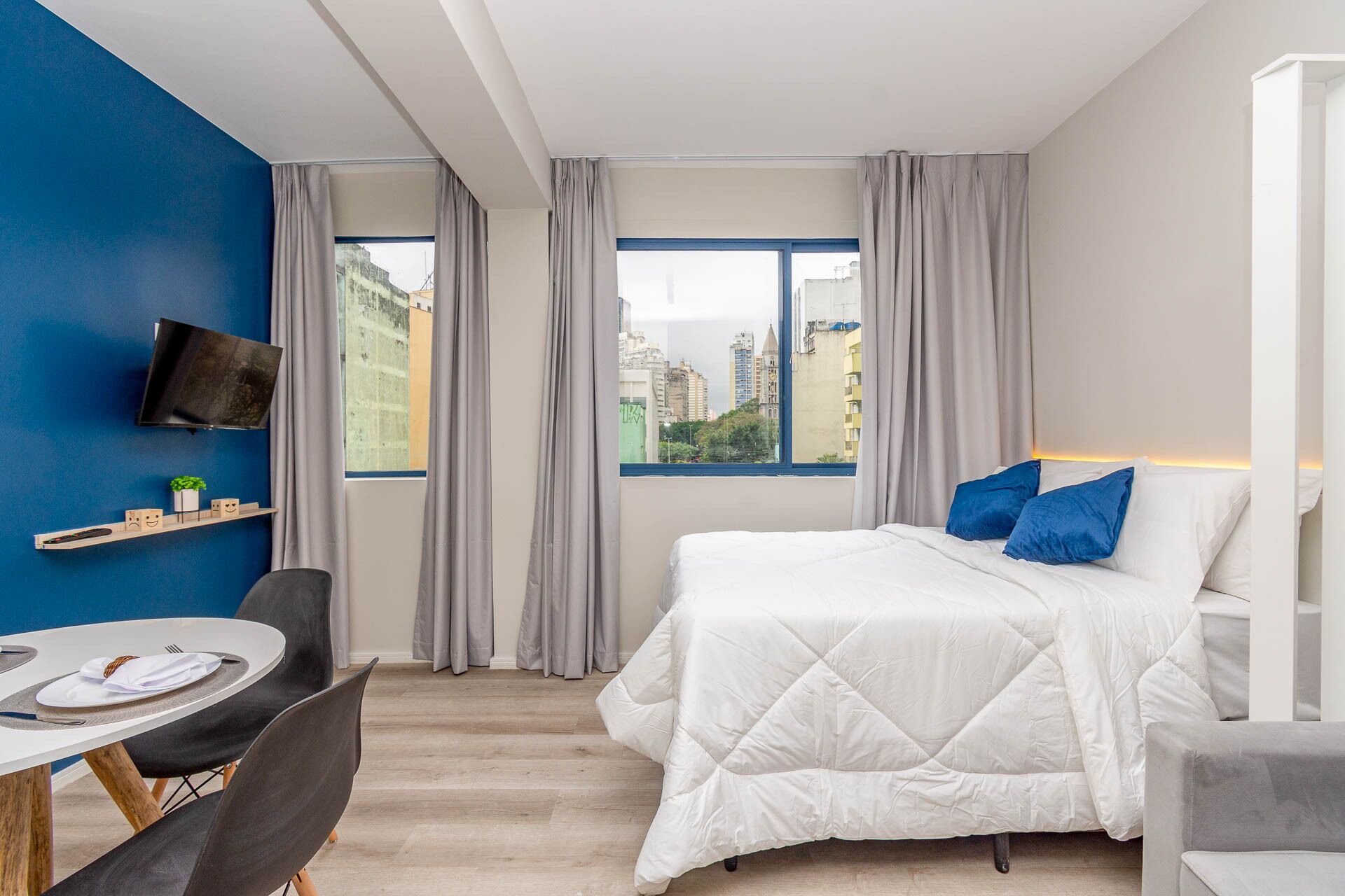 A hotel room with a bed, a TV mounted on the wall, a dining table with chairs, and a view of the city through large windows.