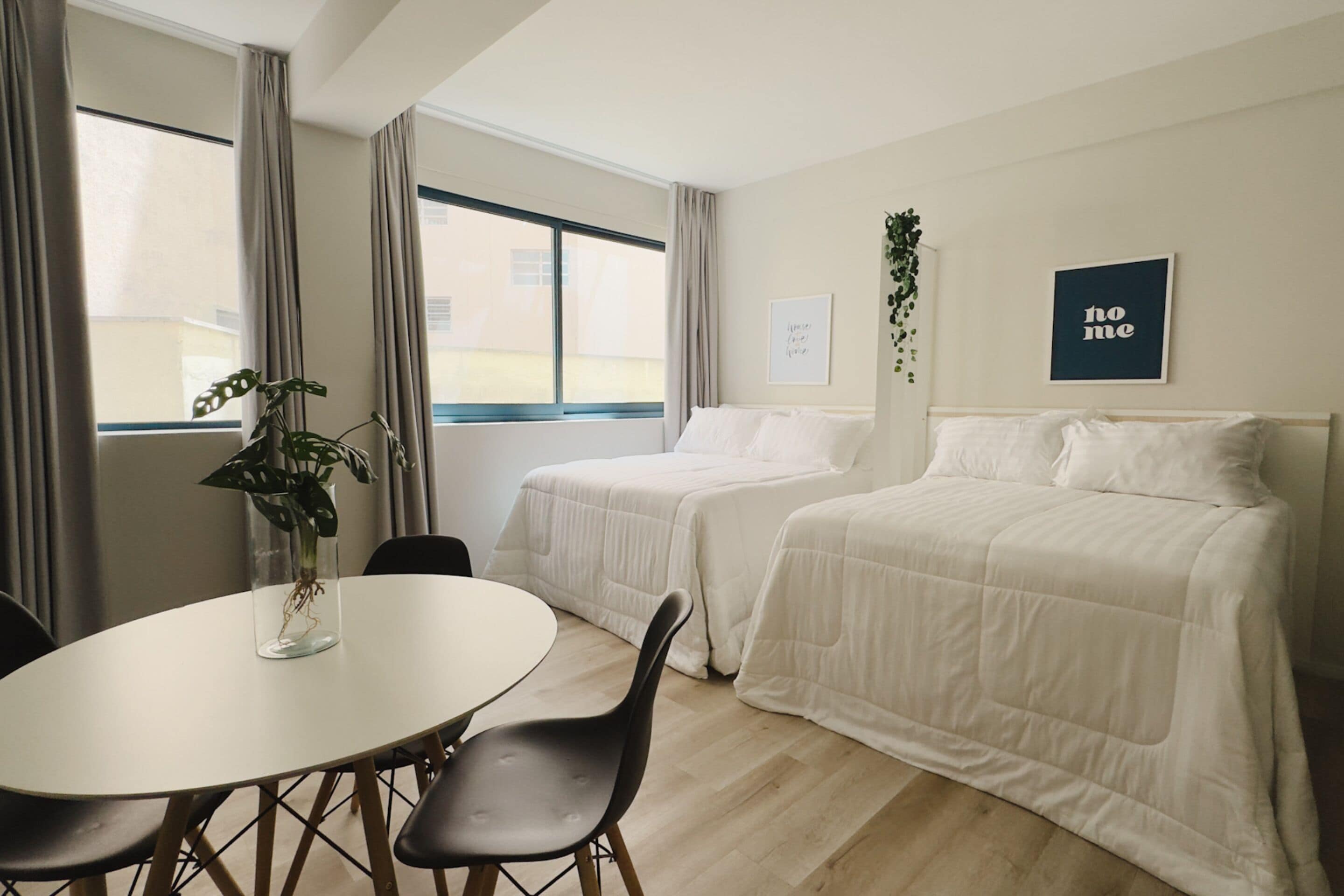 Quarto de hotel com duas camas, uma mesa de jantar, cadeiras, um vaso com planta e um letreiro com a palavra 'home'.