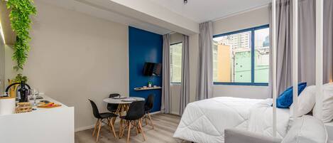 A modern hotel room with a bed, a desk with a chair, a TV, and a window with curtains.
