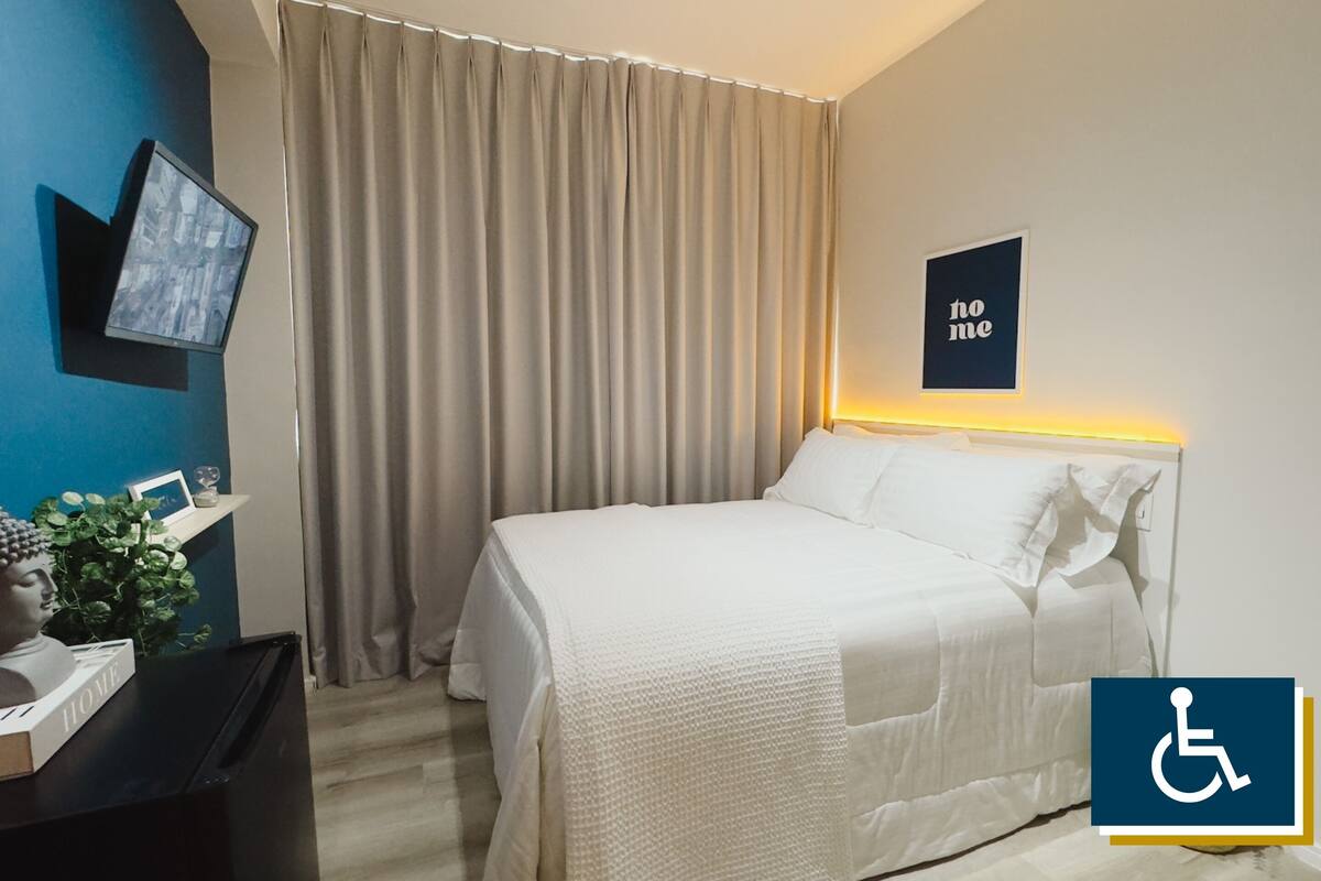Une chambre d’hôtel équipée d’un lit, de rideaux, d’un téléviseur, d’un tableau accroché au mur et d’un panneau indiquant qu’elle est accessible aux personnes en fauteuil roulant.