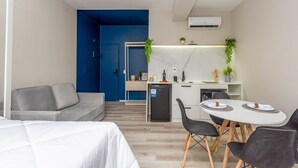Un espacio habitable moderno y funcional con sofá gris, una kitchenette pequeña, zona de comedor con mesa y sillas, y cama.
