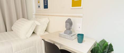 A hotel room with a bed, a desk with a bust sculpture, a mug, and framed artwork on the wall.