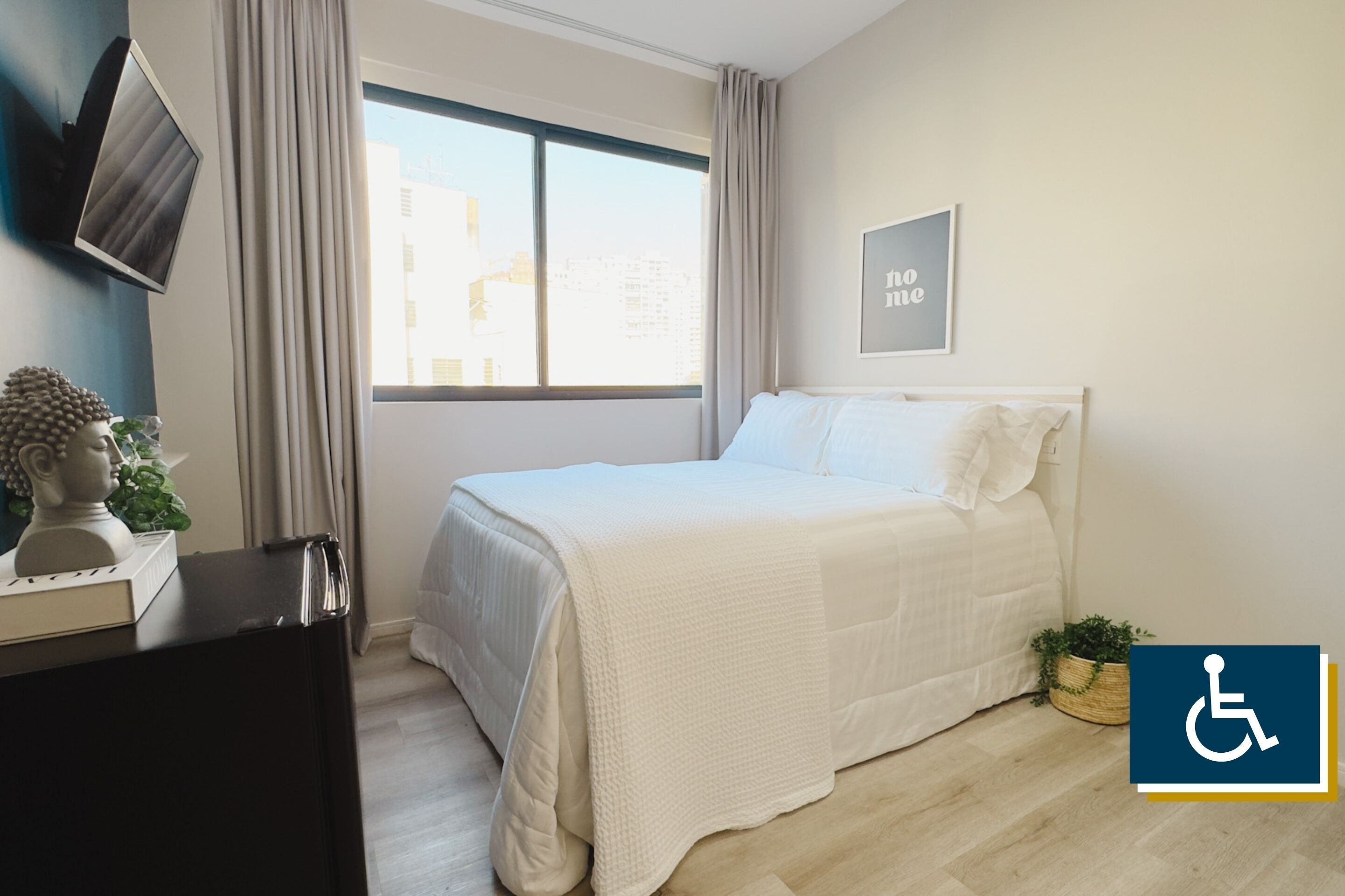 A hotel room with a bed, a TV mounted on the wall, a window with curtains, and a wheelchair accessibility sign.