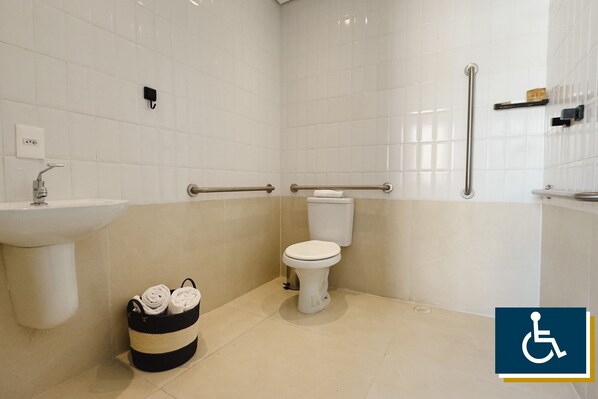 Une salle de bain avec des carreaux blancs, un lavabo, des toilettes, une barre d’appui et une poubelle.