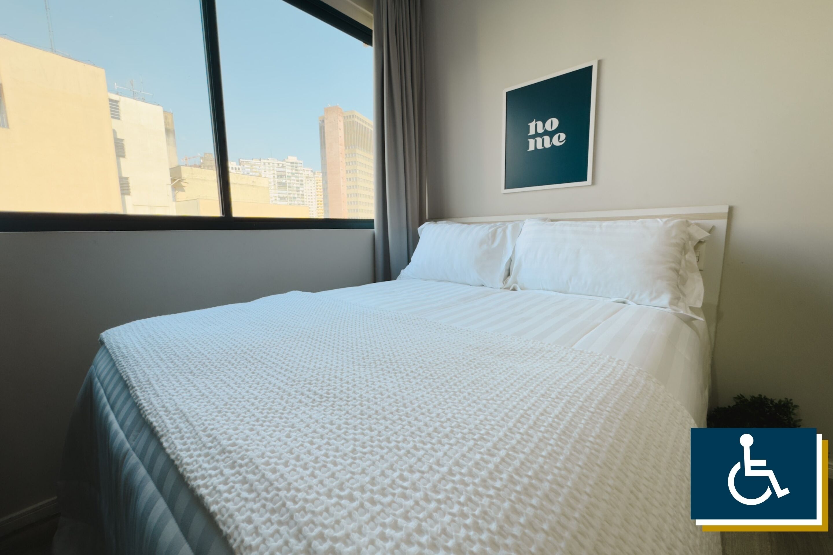 A hotel room with a large window, a bed with white bedding, a framed sign on the wall, and a wheelchair accessibility symbol.