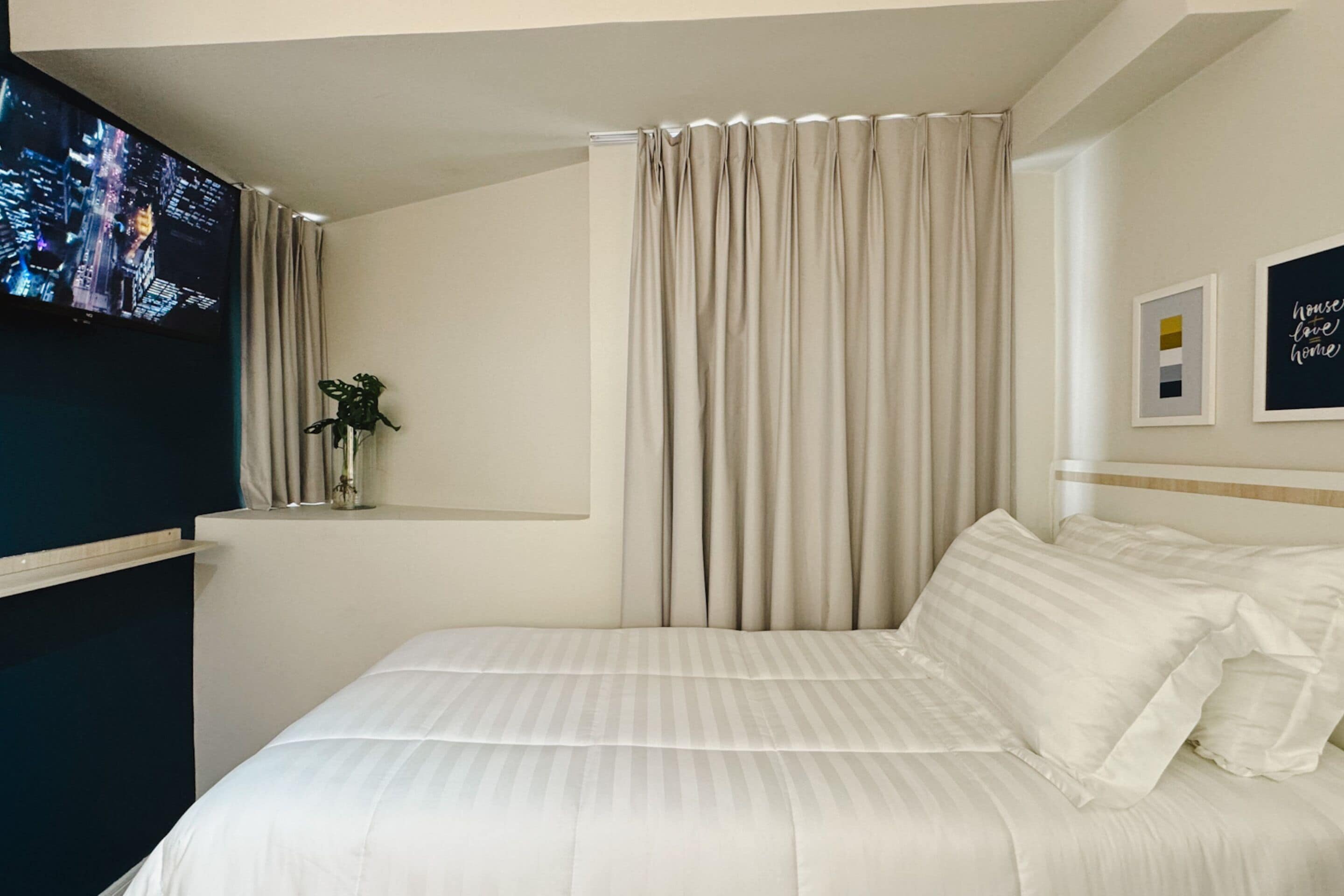 A hotel room with a bed, a TV mounted on the wall, a plant on a shelf, and curtains.