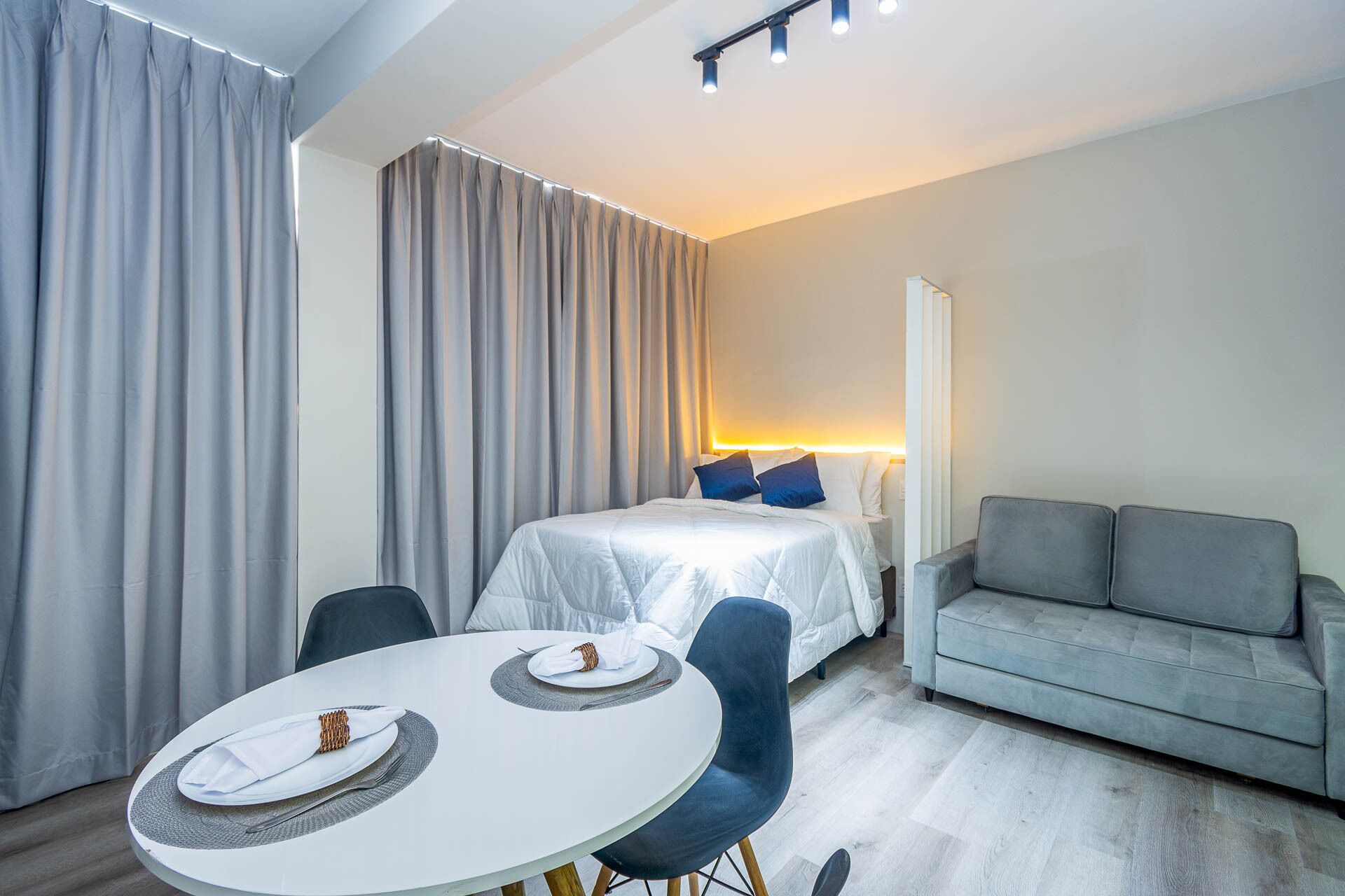 Ein modernes Hotelzimmer mit einem Bett, einem grauen Sofa, einem runden Esstisch mit Tellern und einem großen Fenster mit Vorhängen.