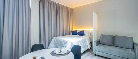 Ein modernes Hotelzimmer mit einem Bett, einem grauen Sofa, einem runden Esstisch mit Tellern und einem großen Fenster mit Vorhängen.