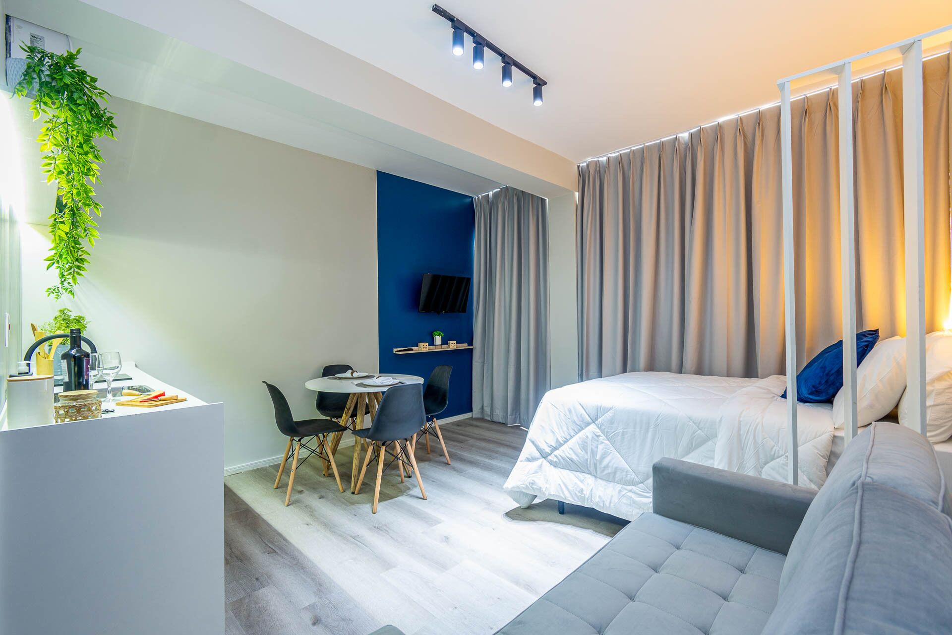 A modern hotel room with a bed, a seating area with a table and chairs, a TV mounted on the wall, and a plant on the left.