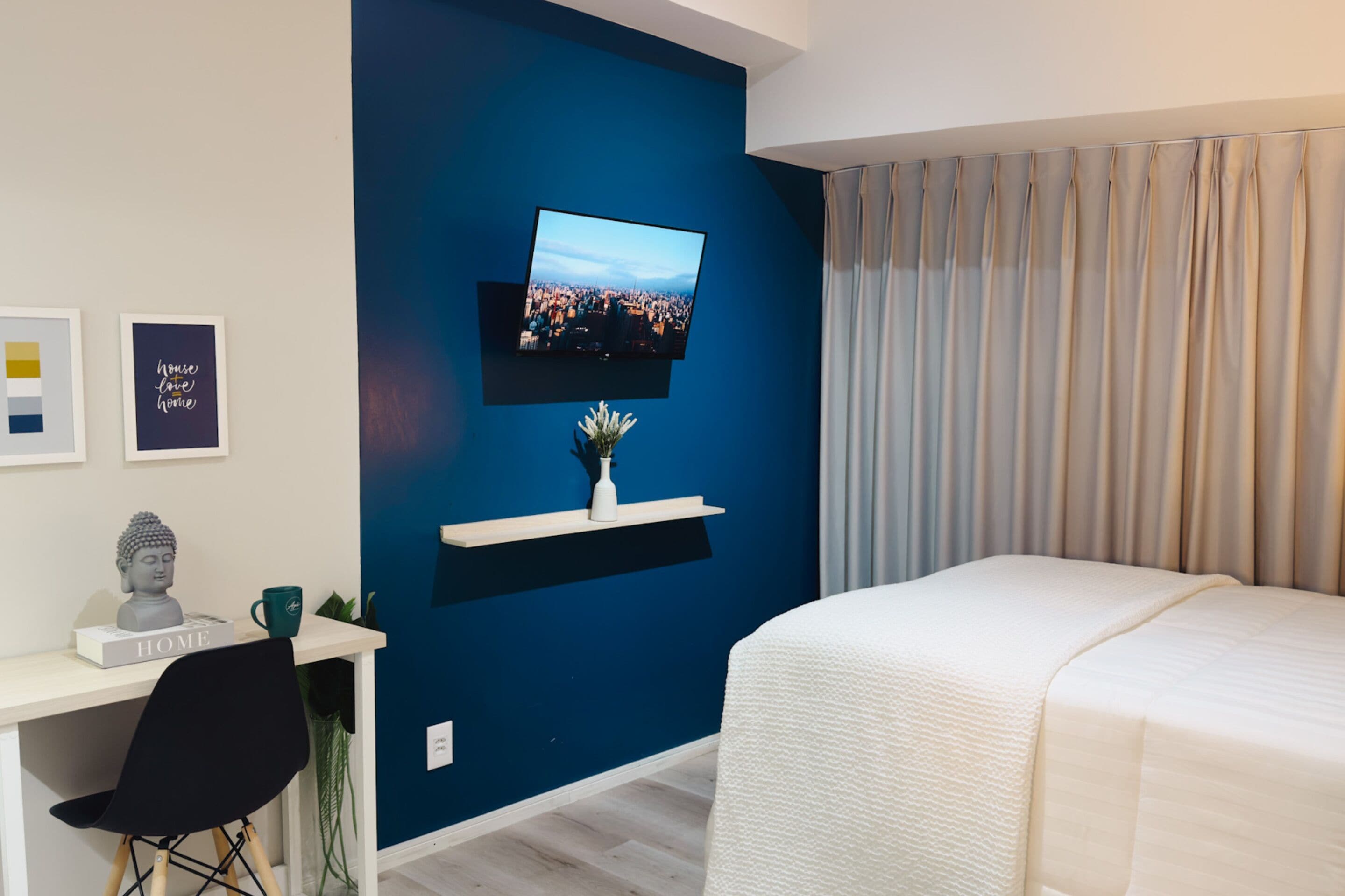 Ein modernes Schlafzimmer mit einem großen Bett, einem Schreibtisch mit Computer, einem an der Wand montierten Fernseher und einem Fenster mit Vorhängen.