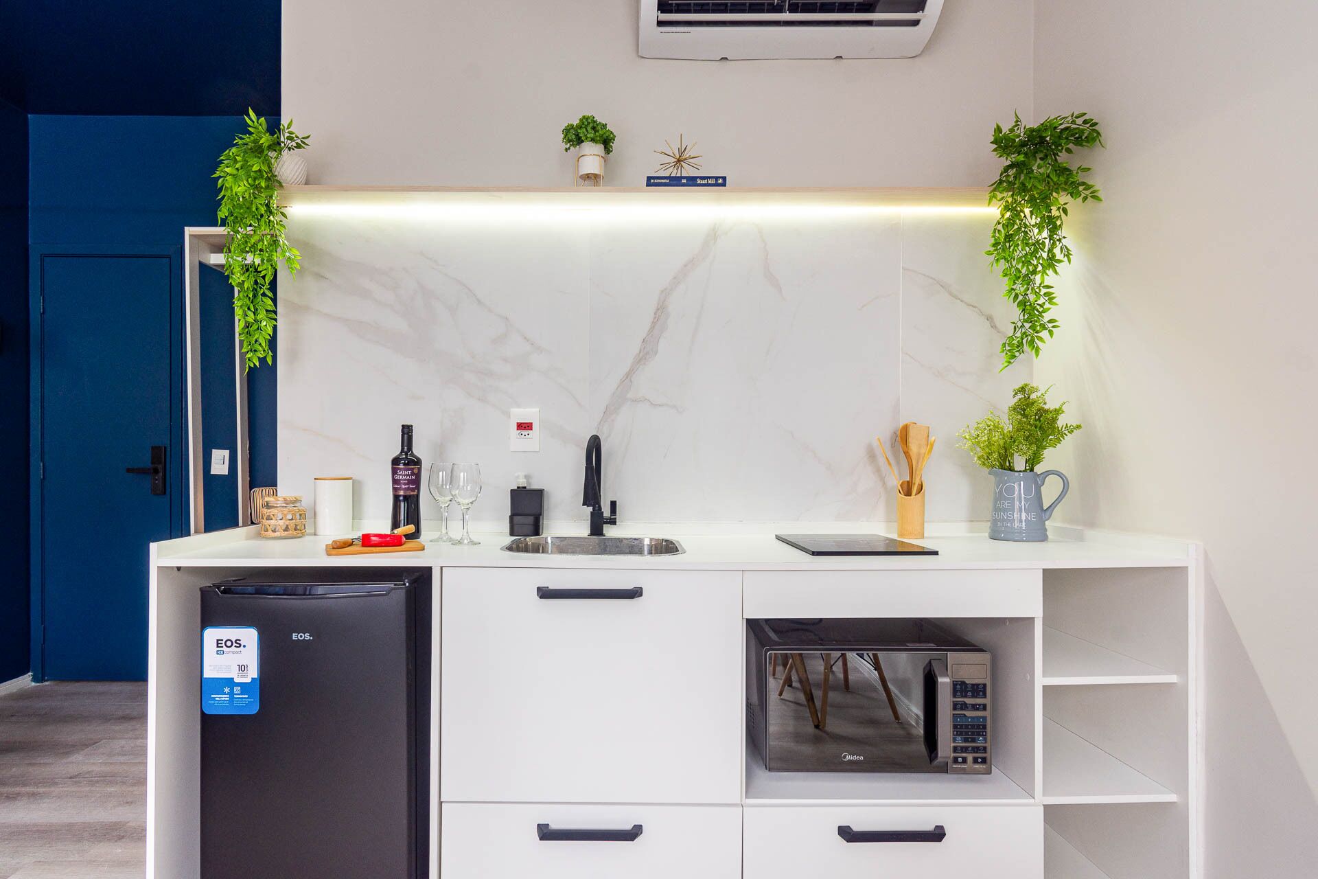 A modern kitchen with a built-in dishwasher, a microwave, and a sink. There are plants on the counter, a book titled 'Street Art', and a sign that says 'YOU ARE MY SUNSHINE'.