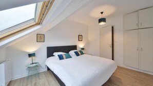 Een slaapkamer met een dakraam, een bed met witte beddengoed en blauwe kussens, een nachtkastje met een lamp en een kledingkast.