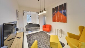Een moderne woonkamer met een bank, fauteuils en een salontafel. Aan de muur hangt een schilderij en een raam zorgt voor natuurlijk licht.