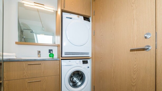A compact laundry room with a front-loading washing machine and dryer, wooden cabinets, a mirror, and a towel rack. - Hrimland - Luxury Apartment for 2 people (Akureyri)