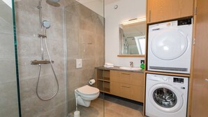 A modern bathroom with a glass-enclosed shower, a toilet, a sink, and a stacked washer and dryer. - Hrimland - Luxury Apartment for 2 people (Akureyri)