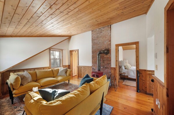 Una habitación acogedora tipo loft con techo inclinado, un sofá amarillo, chimenea de ladrillo y una cama con cabecera de madera.