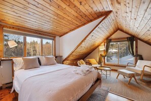 Un dormitorio acogedor con una cama grande, un escritorio y una silla. Hay ventanas con cortinas y vistas a árboles.