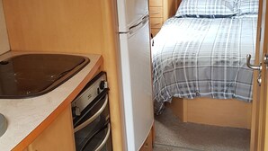 A compact RV interior with a kitchenette, a refrigerator, a microwave, a stove, and a bed with a plaid bedspread. - Lovely Caravan Near Notts and Newark (Newark)