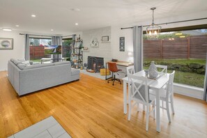 Una sala de estar moderna con un sofá gris, piso de madera, chimenea, zona de comedor con sillas blancas y ventanas amplias con vistas al exterior.