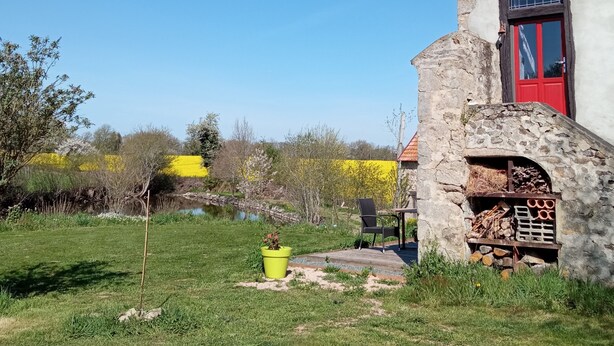 Una casa de piedra rústica con una puerta roja y una chimenea, rodeada por un patio cubierto de hierba y un campo amarillo al fondo.