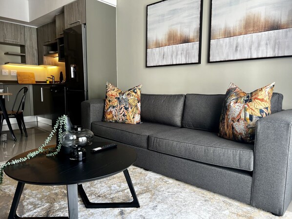 A modern living room with a grey sofa, two patterned throw pillows, a black coffee table with a teapot and remote, and a kitchen in the background.