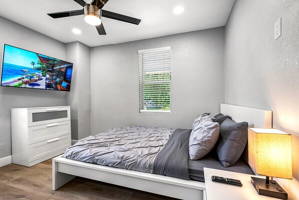 A bedroom with a bed, a TV mounted on the wall, a ceiling fan, a window with blinds, and a bedside table with a lamp. - Remodeled 3BR, 6 Min to Busch Gardens | Fenced Yard, Monthly Stays Welcome (Tampa)
