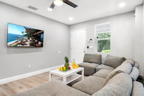A modern living room with a large flat-screen TV, a white coffee table with fruit, a grey sofa, and a window with blinds. - Remodeled 3BR, 6 Min to Busch Gardens | Fenced Yard, Monthly Stays Welcome (Tampa)