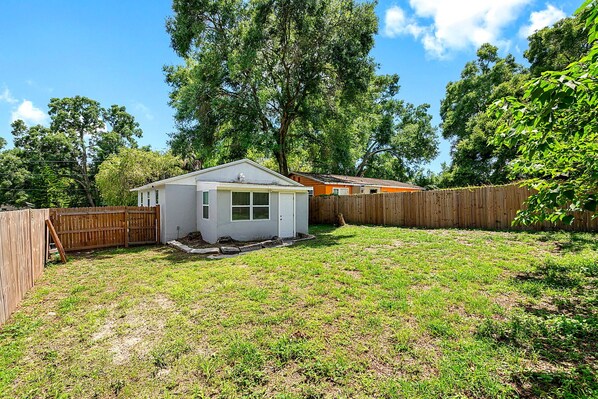 A small, single-story house with a fenced backyard, surrounded by trees and a clear sky. - Remodeled 3BR, 6 Min to Busch Gardens | Fenced Yard, Monthly Stays Welcome (Tampa)