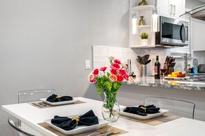 A modern kitchen with white cabinetry, a central island with a vase of flowers, and a dining area set for four with black napkins tied with gold ribbons. - Remodeled 3BR, 6 Min to Busch Gardens | Fenced Yard, Monthly Stays Welcome (Tampa)